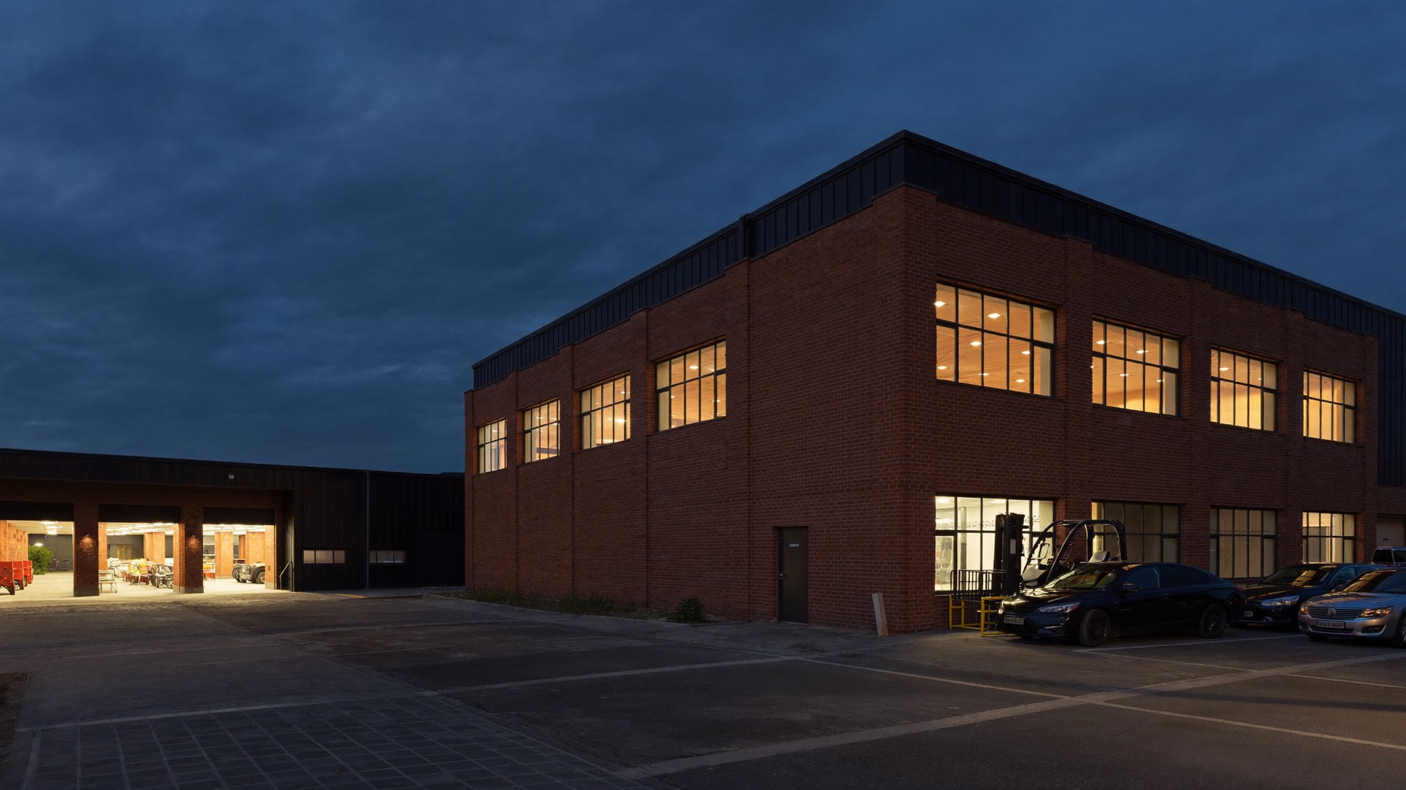 Familiengeführter Mittelständler am Abend: Produktionsgebäude aus Backstein und Stahl mit erleuchteten Fenstern, davor der Firmenhof