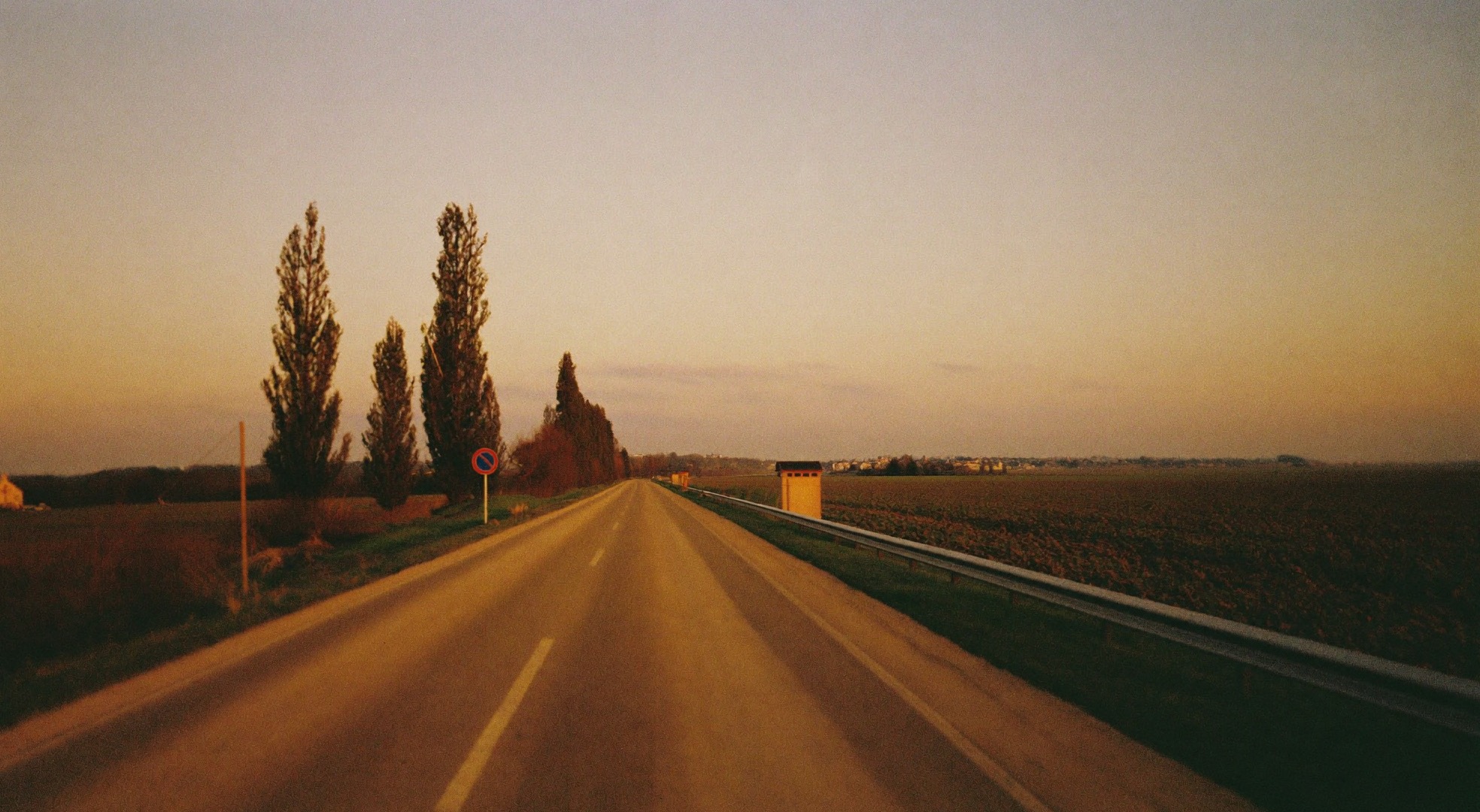 Leere Landstraße im Abendlicht, die zwischen Feldern auf einen Ort am Horizont zuläuft