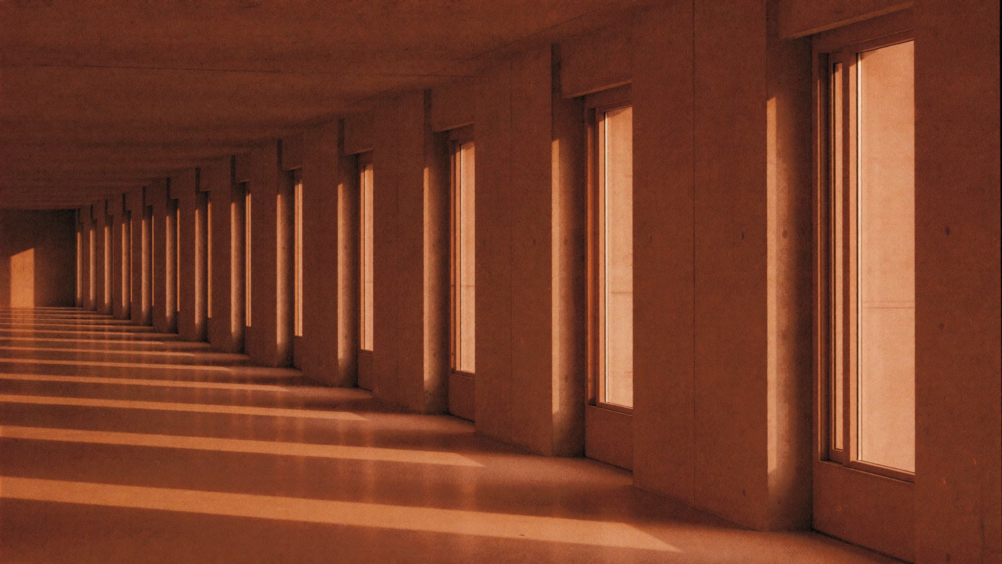 Reihe gleichmäßiger Fensterachsen in einem modernen Gebäude, durch die das Abendlicht in gleichen Abständen auf den Boden fällt